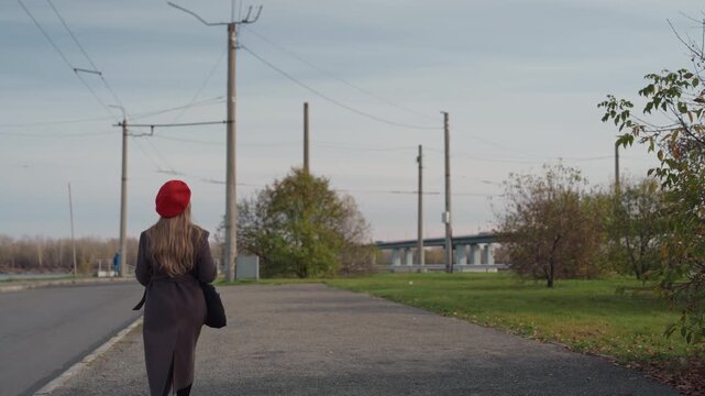 Caucasian woman walking on rural road in red beret and long coat, carrying black bag, late autumn atmosphere, overcast sky, utility poles and bridge in distance, gravel shoulder and sparse trees,