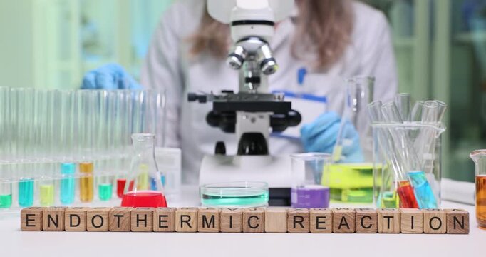 Woman laboratory worker observes chemical reaction through microscope. Wooden cubes on laboratory table form phrase Endothermic Reaction by glassware