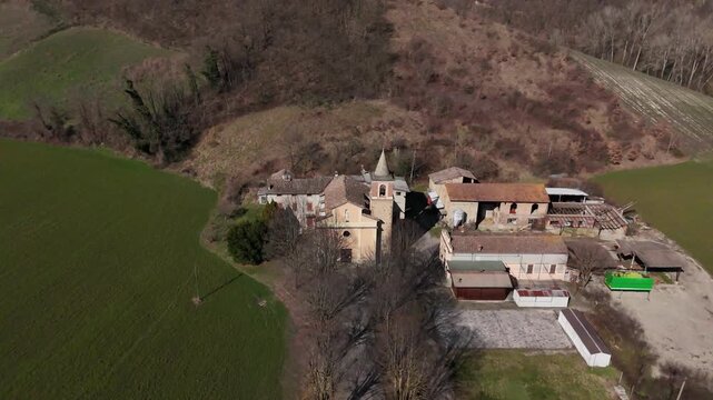 Sant'Ilario Vescovo church and medieval village cluster in Chiavenna Rocchetta, Italy, surrounded by bare trees, green fields, and rolling hills in a rural setting