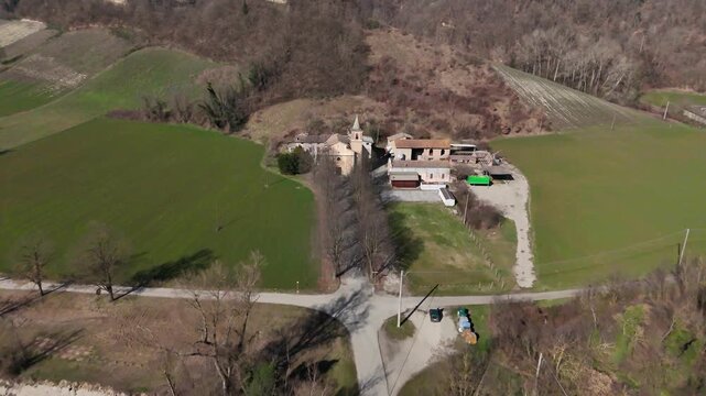 Sant'Ilario Vescovo church and medieval village cluster in Chiavenna Rocchetta, Italy, surrounded by bare trees, green fields, and rolling hills in a rural setting