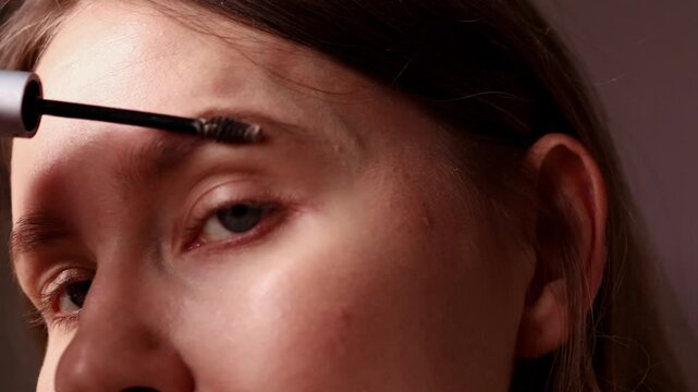 A young woman holds a tube of eyebrow gel and uses a small spoolie brush applicator to style and tint her eyebrows. Close up of facial makeup and brow grooming routine