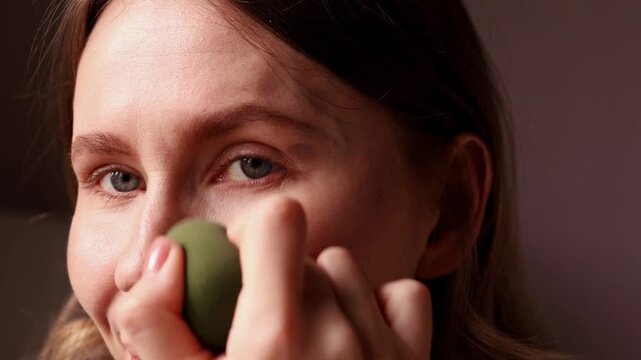 A young woman uses a dark egg-shaped cosmetic sponge to blend foundation or concealer on her face. Close up of natural makeup routine and skin application