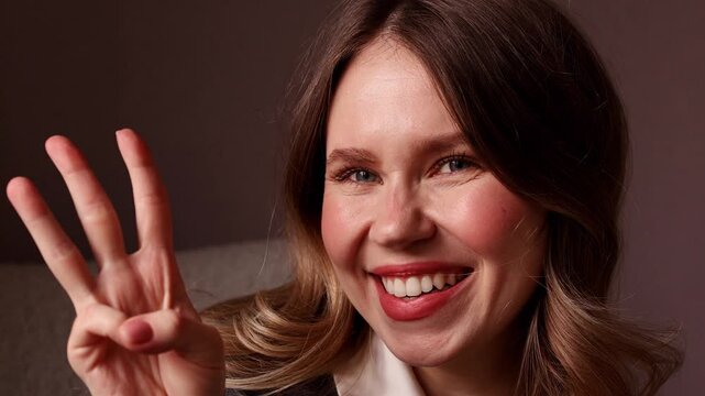A cheerful young woman looks at the camera with a bright smile, progressively showing one, two, and three fingers as she counts against a dark background