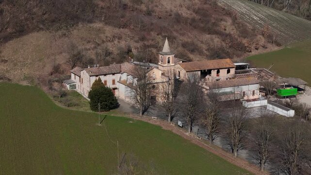 Sant'Ilario Vescovo church and medieval village cluster in Chiavenna Rocchetta, Italy, surrounded by bare trees, green fields, and rolling hills in a rural setting