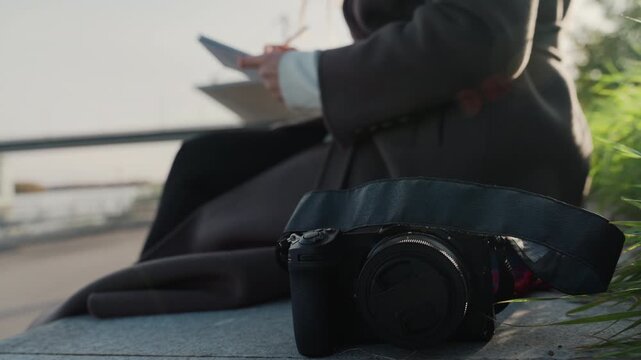 Sketchbook on riverside bench, camera foreground, urban sketcher in coat drawing city skyline at golden hour, shallow depth of field with blurred bridge and soft bokeh, tactile camera gear