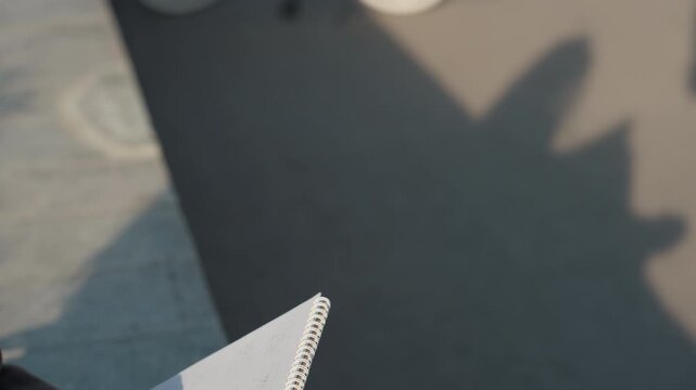 Notebook held casting shadow on pavement, closeup of spiral notebook edge and visible palm, textured asphalt grain, warm afternoon sunlight, contemplative street sketching, journaliststyle notetaking,