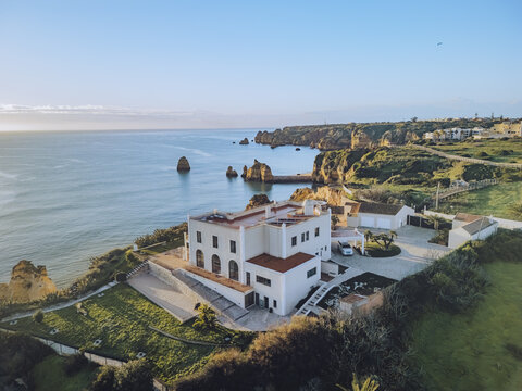 Aerial view of a grand white building with terracotta roof tiles standing proudly on a green cliff overlooking the azure sea and rugged coastline, Lagos, Faro District, Portugal.
