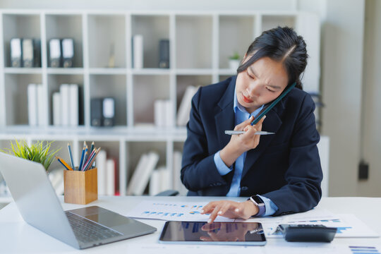 Young asian businesswoman managing multiple tasks, talking on a smartphone while simultaneously using a digital tablet to analyze data and reviewing financial reports in a modern office environment