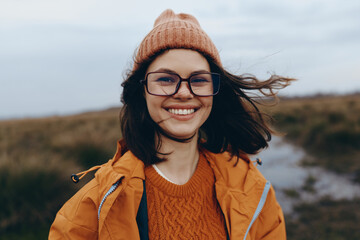 Smiling woman in orange jacket and knit hat stands outdoors on a windy day, natural landscape in...