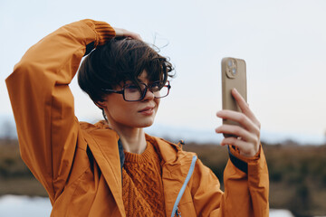 Young woman lifestyle taking selfie outdoors wearing glasses and orange jacket with hand in hair....