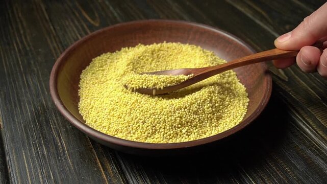 Hand scooping yellow couscous with wooden spoon from a brown bowl on a rustic wooden table, showcasing the texture and movement of the grains
