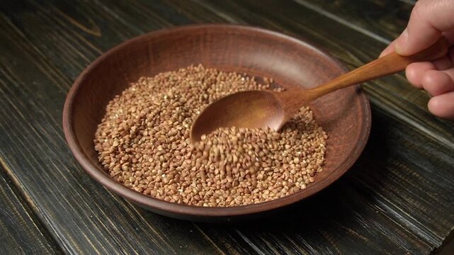 Hand scoops buckwheat from a wooden bowl with a wooden spoon, capturing the continuous motion and texture of the seeds against a dark wooden surface in a cozy kitchen environment