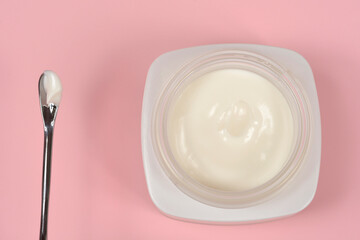 A top view of an open white jar filled with smooth face cream and a metal cosmetic spatula with a drop of cream on a solid pink background