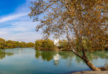 Obraz premium White lighthouse tower reflecting in a calm river with turquoise water. Scenic landscape with autumn trees, yellow leaves, and blue sky in Manavgat, Antalya province, Turkey. Sunny November day.