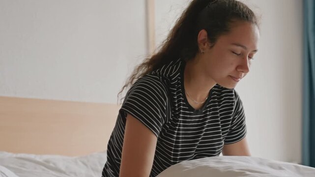 Young woman in hotel bed waking slowly, groggy movements, massaging temple and wiping eyes, daylight through curtains, bedside lamp, headboard and crisp sheets set scene of sleepless night