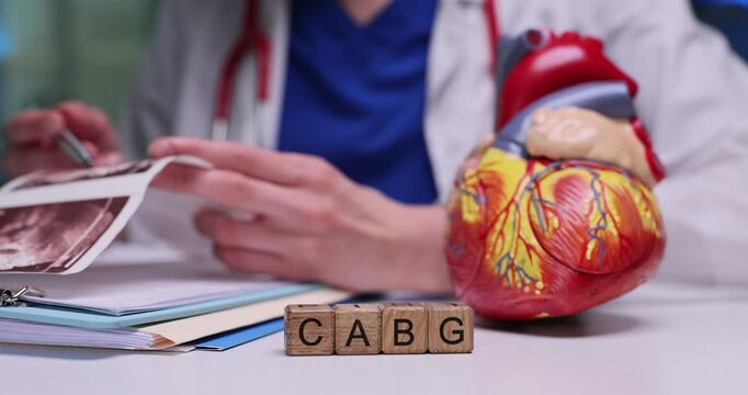 Woman cardiologist reviews detailed heart ultrasound scans of patient. Wooden cubes form abbreviation CABG near anatomical heart model on table