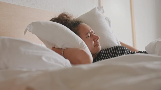 Sleepy hotel guest muffles annoying morning noise with pillow woman in polka dot dress presses pillow over head, flips and buries face in white duvet, soft daylight through neutral curtains, intimate