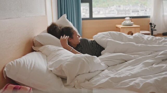 Overwhelmed hotel guest hides under pillow on rumpled bed wide shot shows woman pulling pillow over face beside window with city view, bedside lamp and travel bag suggest stay, daylight floods room,