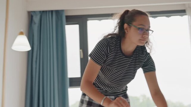 Hotel staff inspecting finished bed, leaning over mattress and adjusting pillows, checking presentation against checklist, calm efficient posture with emphasis on quality and hospitality standards