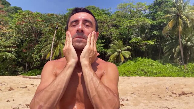 Man applying sunscreen cream on his face while standing on a tropical beach with palm trees. Sun protection, skincare, summer vacation, and healthy outdoor lifestyle concept
