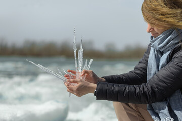 Woman hold sharp transparent ice crystals on frozen river during spring thaw against blurred winter landscape. Concept of seasonal climate change, beauty of nature, human connection with environment.