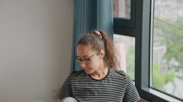 Guest leaning to inspect hotel view of cityscape, attentive traveler peering through windowpane with glasses and striped shirt, quiet discovery moment, urban buildings and wires outside framing