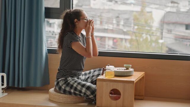 Woman eating hotel breakfast by window, low table with tray and juice, bento and chopsticks on wooden table, sunlight through curtains, cozy seat and city view, relaxed travel guest savoring morning