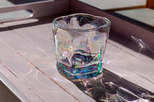 Glass tumbler with iridescent coating on wooden surface. Shimmering light reflections and abstract shadow. Modern drinkware display concept.