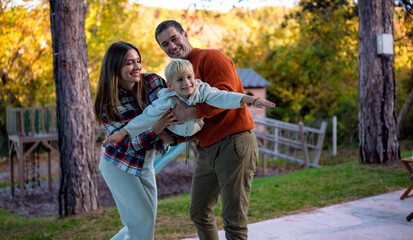 Fototapeta premium Happy family spending quality time together and having fun outside, parents holding their son pretending he is flying in the park during a sunny autumn day