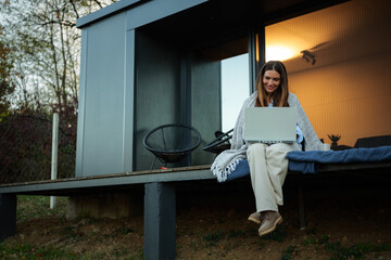 Woman wearing a cozy blanket and smiling, working on a laptop while sitting on the modern house...