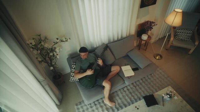 Relaxed Individual Engages In Lively Guitar Session Amidst Scattered Papers And Warm Lighting