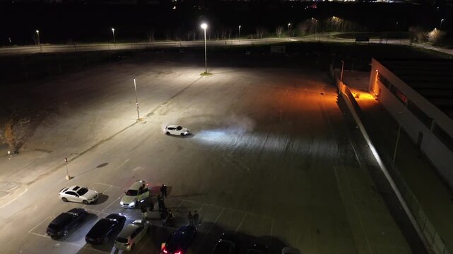 A white car slides into the corner, its paint glowing under the streetlights like fresh snow. The engine roars as the driver snaps the steering wheel, and the rear tires break loose with a sharp screa