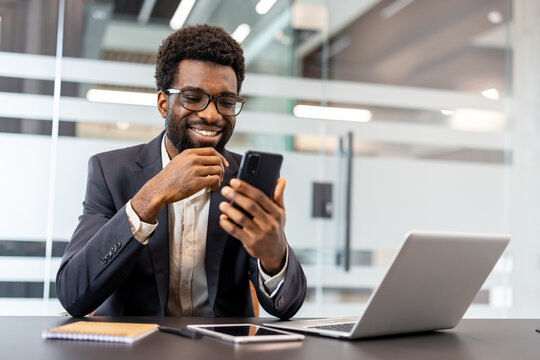 Smiling businessman browsing smartphone at a desk with open laptop in a modern office, looking positive and connected while working remotely and staying productive and professional