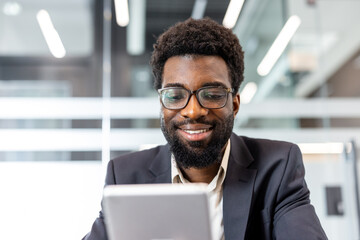 Obraz premium Young man wearing glasses and a business suit confidently working and smiling while using a digital tablet for business communication in a bright contemporary workspace