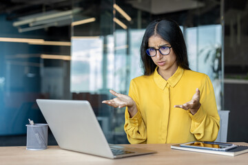 A young Indian businesswoman is sitting at her office desk and enjoying a video call on her laptop