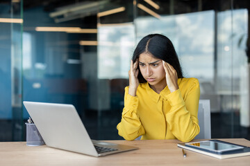 Upset and worried young Indian woman sitting at a desk, looking at a laptop screen and holding her head