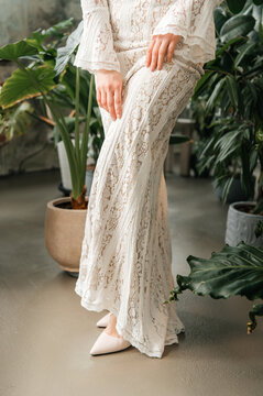 ow angle view of a woman wearing a bohemian style lace dress and beige high heels, posing in a botanical studio or greenhouse with lush indoor plants.