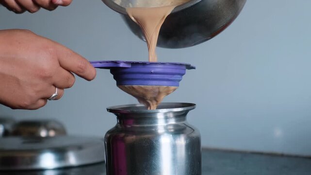 Close-up of hands straining hot Indian masala tea through a purple sieve into a steel pot