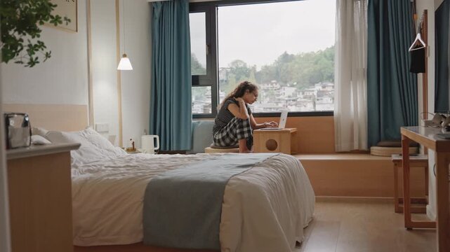 Hotel room writer types at window, solo guest writing on laptop on window seat, soft morning light, tidy bed with blue throw, city view beyond curtains, sipping tea between paragraphs, reflective