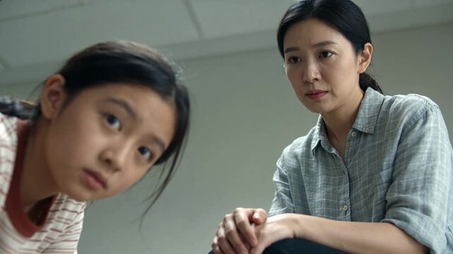 Noticing off-camera, child leaning in striped tee, mother in check shirt clasping knee in clinic