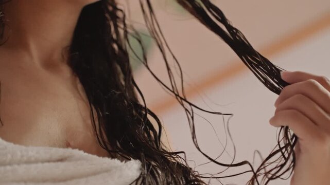 Towel wrapped guest examines wet braids in hotel room, spa guest enjoying calm unwind, fingers twist and separate strands, warm ambient light, soft skin and chest profile, quiet leisure moment