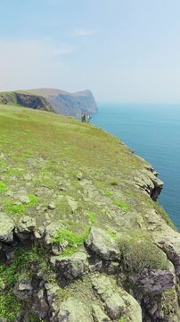 Aerial view of a group of young men and women standing on a high cliff edge overlooking the Jernas shipwreck, Salalah, Oman.