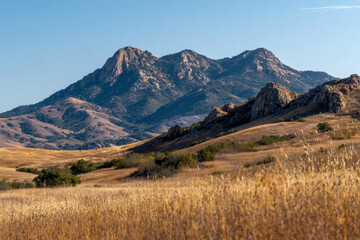 Obraz premium Rolling golden grasslands with rugged rocky hills and towering mountain peaks under clear blue sky in a peaceful natural landscape setting