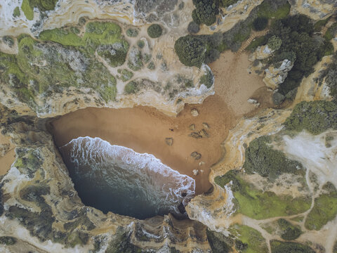Aerial view of a secluded cove with golden sands meeting the turquoise sea, framed by rugged cliffs adorned with patches of green, Albufeira, Faro, Portugal.