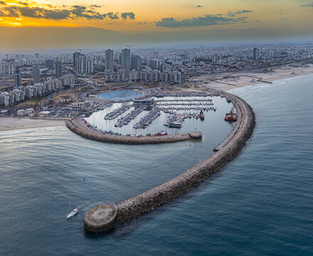 Aerial view of the marina's protective arm embracing a fleet of yachts against the azure sea, juxtaposed with the city skyline, Ashdod, South District, Israel.