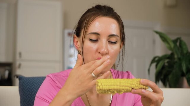 Girl licking fingers while holding corn cob in hand. Woman finishing bite of maize during meal at kitchen table. Lady eating sweetcorn and cleaning fingers after snack