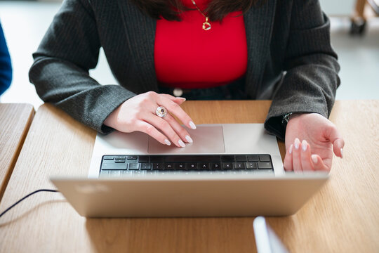 A woman in a suit jacket and red top uses a laptop, her hands resting on the keyboard and trackpad.