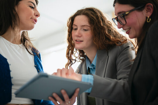 Three young women collaborate on a digital tablet, discussing ideas and sharing information in a bright, modern setting.