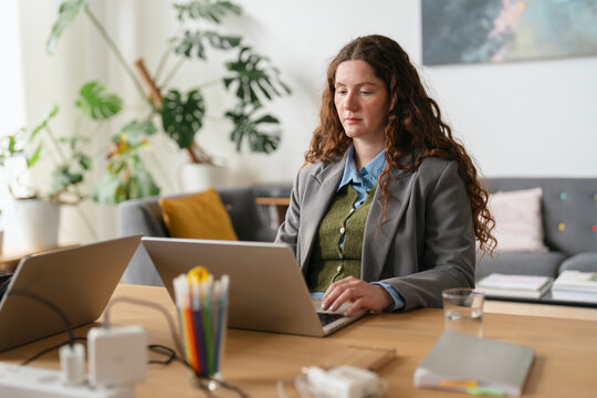 A woman with curly brown hair works on her laptop at a desk, surrounded by plants and office supplies.