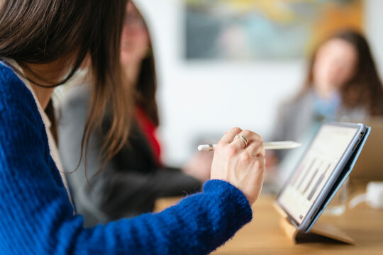 A woman in a blue sweater uses a stylus to interact with a tablet displaying a bar graph during a business meeting.
